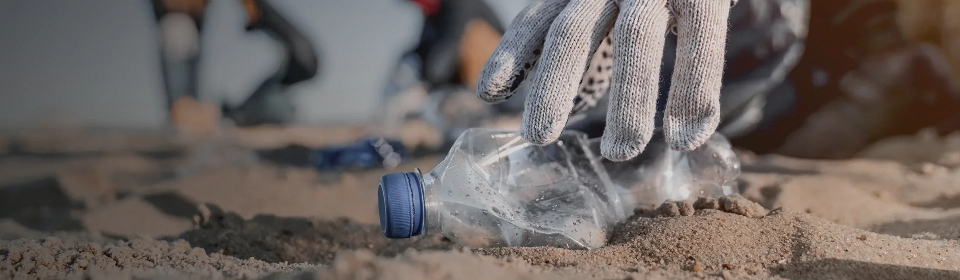 Ręka w rękawicy podnosi plastikową butelkę z piasku, sugerując sprzątanie w plenerze